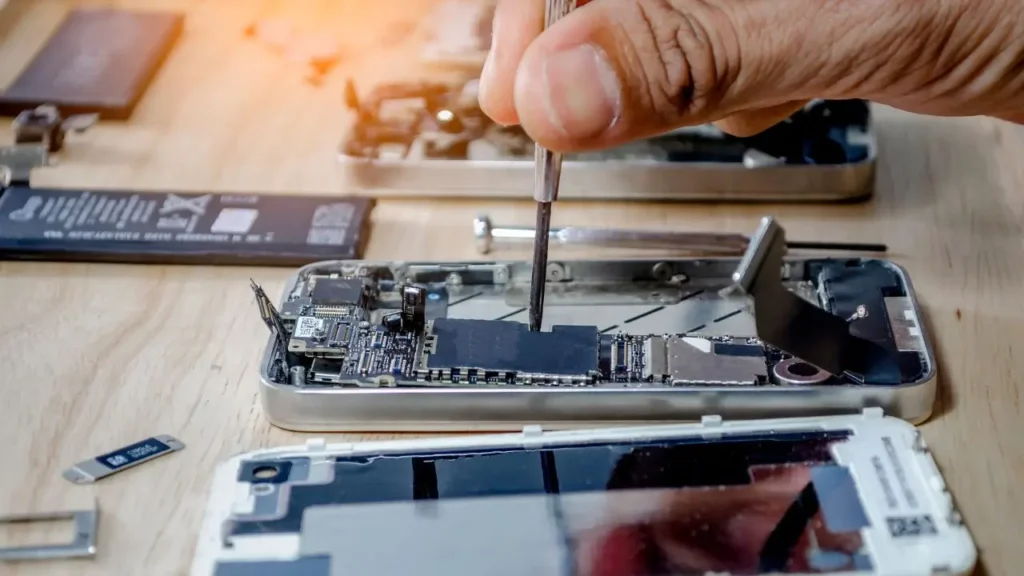 A close-up view shows a person's hands carefully repairing a disassembled smartphone, with various electronic components and a battery laid out on a wooden surface. Location: Toronto, Canada